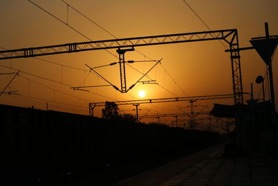 Silhouette electricity pylons by railroad tracks against sky during sunset