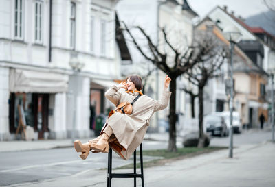 Smiling woman in a beige trench coat enjoys a whimsical moment on a street chair