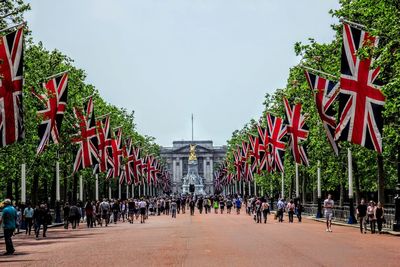 Crowd amidst british flag leading towards buckingham palace against sky on sunny day