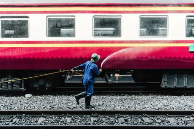 Side view of faceless male worker in blue uniform and hat pouring water from hose in red train on railroad station