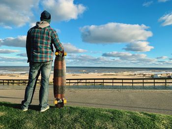 Rear view of man holding skateboard while looking at sea against sky