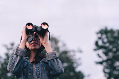 Young woman looking through binoculars while standing against sky