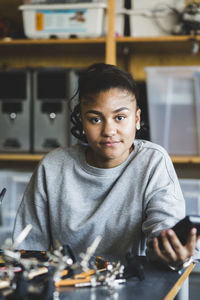 Portrait of confident female teenage student sitting with science project at desk in classroom at high school