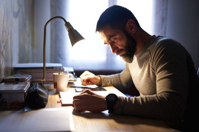 Young white man with a neat beard studying and looking at the phone at his desk