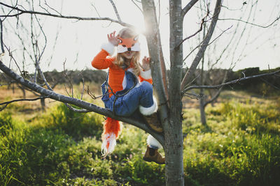 Portrait of girl in fox costume sitting on bare tree against sky during halloween at park