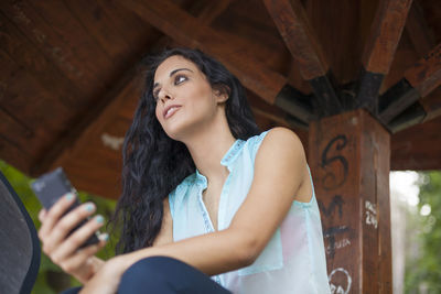 Low angle view of cheerful young woman using phone while sitting in gazebo