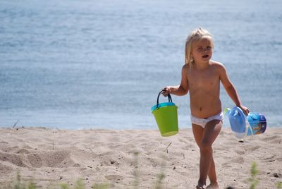 Shirtless girl carrying buckets while walking on sand at beach during sunny day