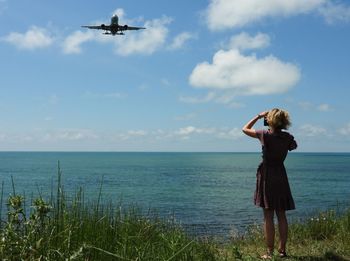 Woman standing in sea against sky and airplane