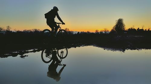 Silhouette man with bicycle by lake against sky during sunset