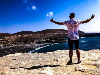 Rear view of man with arms outstretched standing on rock by sea against sky