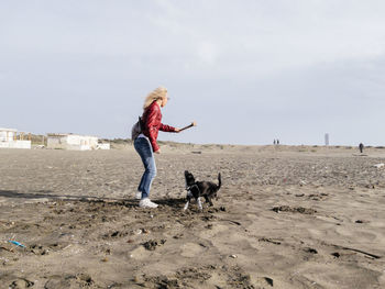 Full length of woman playing with dog on sand at beach against sky