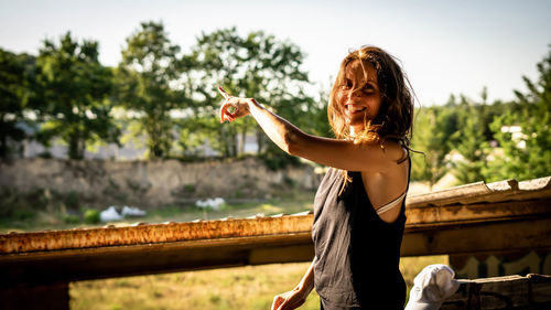 Portrait of young woman with arms raised standing against railing