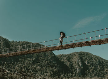 Low angle view of young woman standing on bridge by mountains against sky