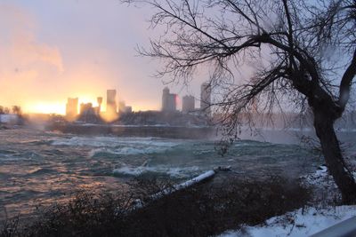 Frozen river by buildings against sky during sunset