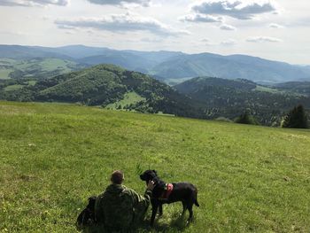 Rear view of man sitting with dogs on grassy field against mountains