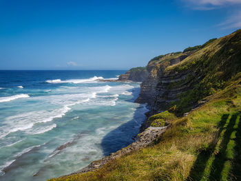 Scenic view of sea with waves and green cliffs against blue sky