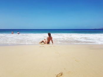 Rear view of woman sitting at sandy beach against blue sky