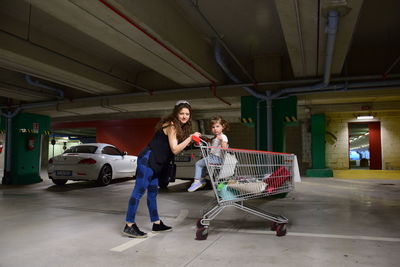 Portrait of mother with daughter sitting in shopping cart at parking lot