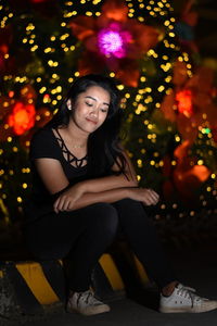 Portrait of young woman sitting on illuminated christmas tree