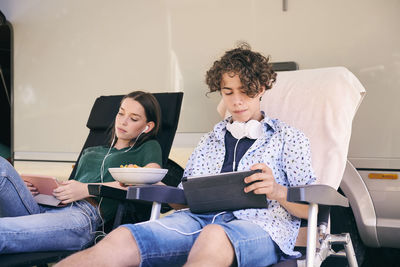 Siblings using digital tablet while enjoying snacks against camper van