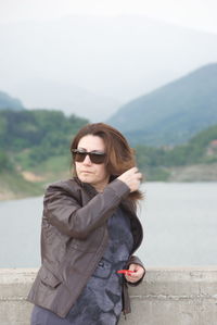 Portrait of woman wearing sunglasses standing by retaining wall against lake