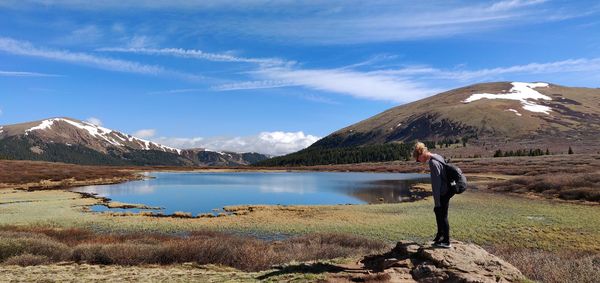 Side view of teenage girl with backpack standing on rock by lake against blue sky during sunny day