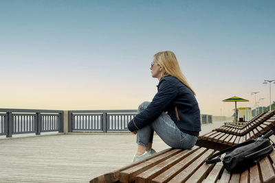 Blonde woman on an empty city embankment rests sitting on a sun lounger and looks into the distance.