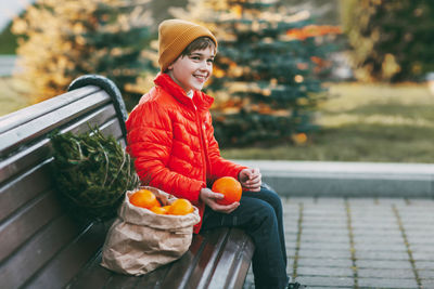 A boy in a bright orange jacket and a fashionable yellow hat sits on a bench, laughs, throws oranges 