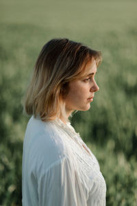 Calm young female dressed in old fashioned blouse and skirt standing alone among tall green grass in cloudy summer day in countryside