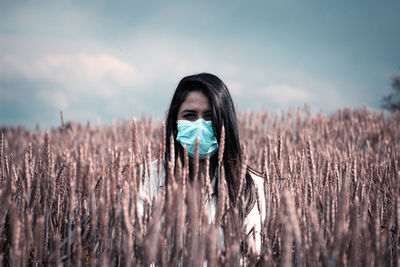 Portrait of young woman wearing face standing amidst plants against sky