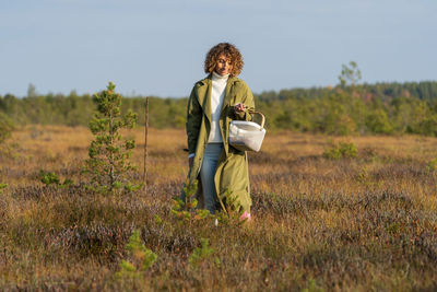 Nature autumn activities. young woman searching cranberries on swamp holding basket happy smiling
