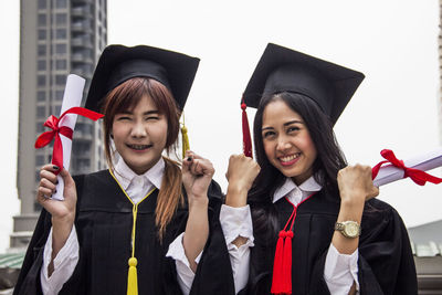 Cheerful young women holding degree certificates standing against sky