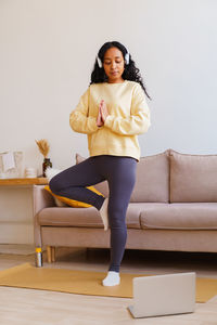 African-american female in headphones standing in tree pose yoga while following tutorial on laptop