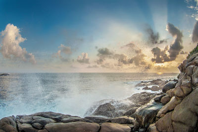 Waves splashing on rocks at beach against sky during sunset