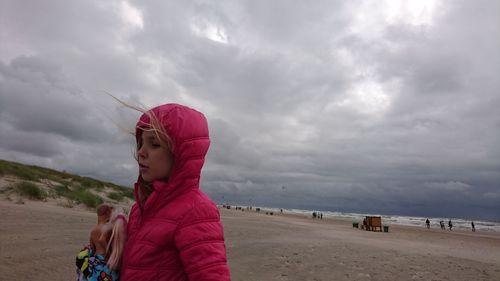 Little girl holding dolls at beach against cloudy sky
