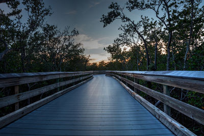 Footbridge amidst trees against sky during sunset