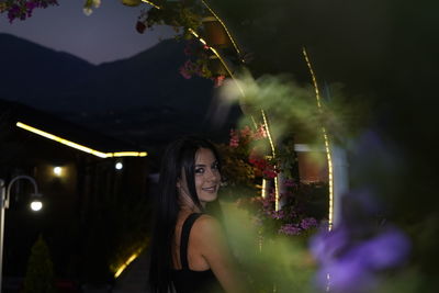 Portrait of young woman standing by illuminated flowering plants
