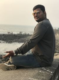 Side view of man sitting on retaining wall while practicing yoga against sea