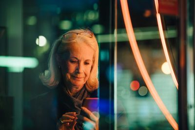 Wrinkled woman using smart phone while standing in city at night