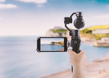 Cropped hand of woman photographing sea through mobile phone in monopod at beach