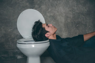 Young man sleeping on toilet bowl in bathroom at home