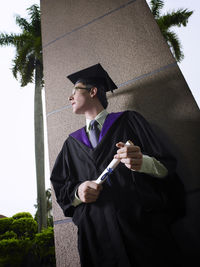 Low angle view of university student wearing graduation down against column