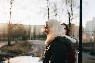 Side view of smiling young muslim woman with female friend walking in city during sunset