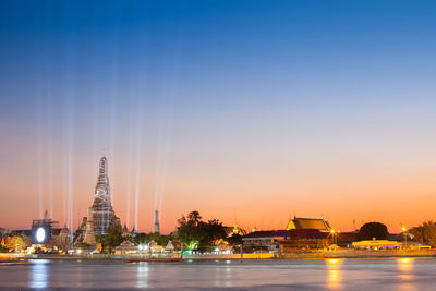 Illuminated buildings by river against sky during sunset