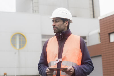 Man wearing reflective vest and hard hat holding tablet looking around