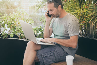 Man using mobile phone and laptop while sitting on seat by plants