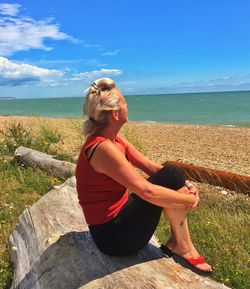 Woman sitting on log at beach against blue sky