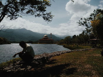 Rear view of woman sitting by river against sky