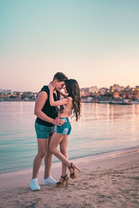 Young couple standing on beach against sky during sunset