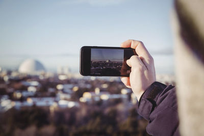 Cropped image of man photographing cityscape through mobile phone during winter